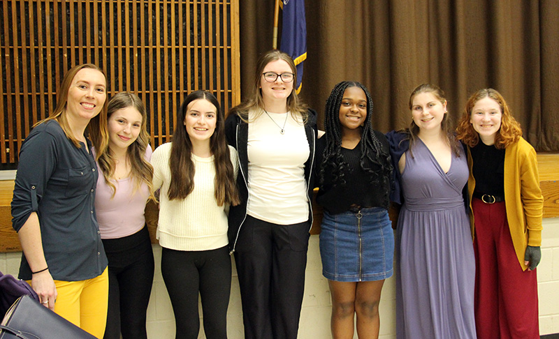 A group of six young women stand with a woman on the left in front of a stage. All are smiling