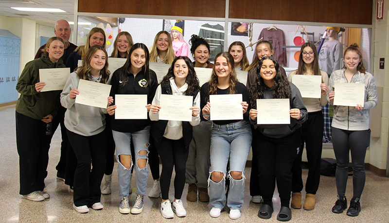 Fourteen high school girls stand together smiling. They are holding up certificates they received. With them is a man standing in the back.