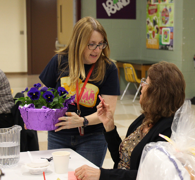 A woman with long blonde hair and glasses places a purple plant on a table in front of a woman who is talking to her.