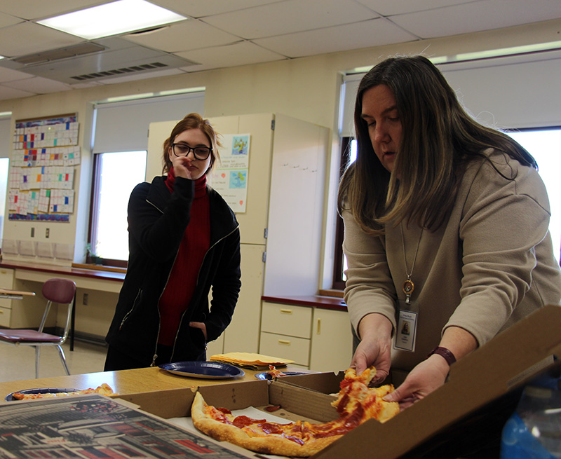 A woman takes a piece of pizza out of a box while a young woman waits for it.
