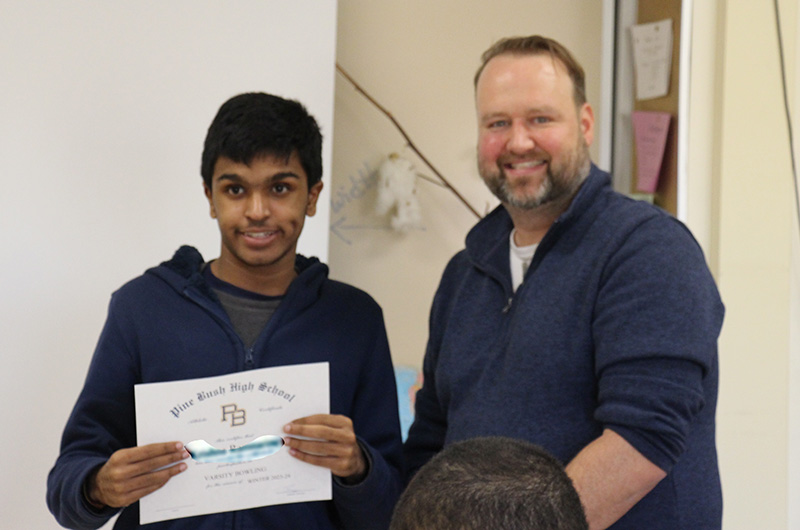 A young man holds a certificate and smiles next to a man with dark hair and a mustache.