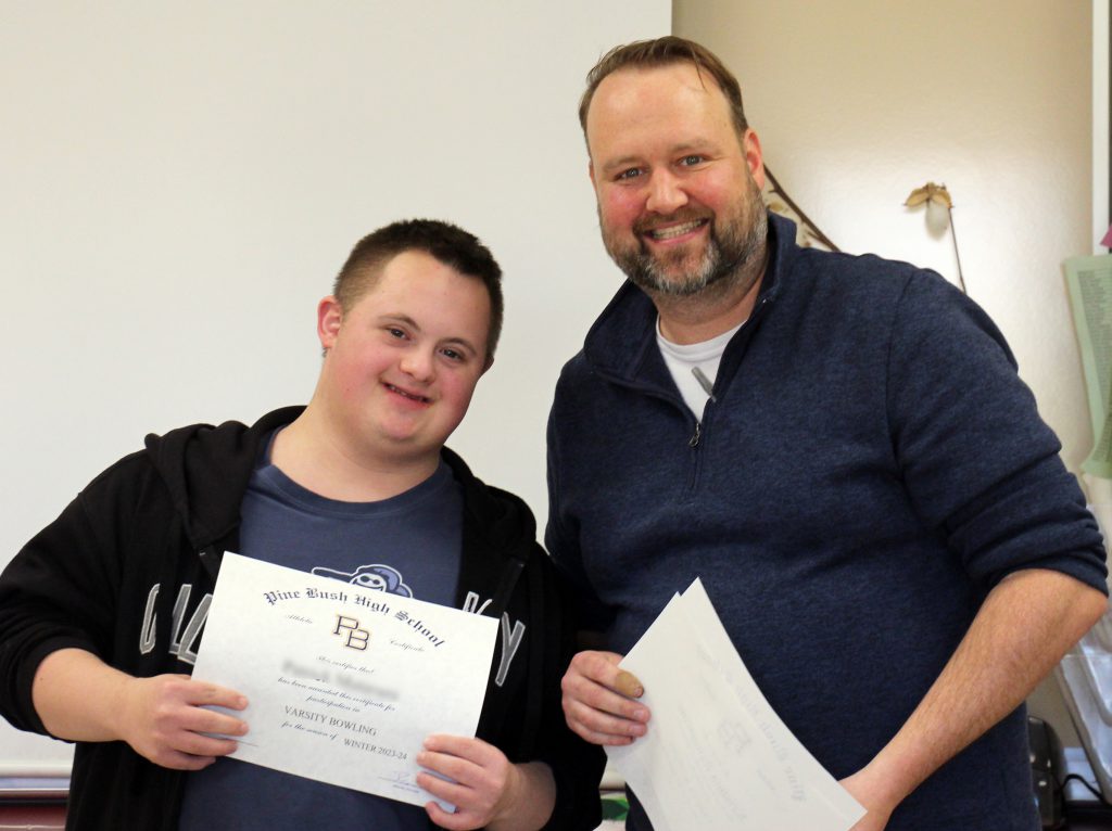 A young man with short dark hair holds a certificate and is standing next to a man smiling, with short dark hair and a mustache.