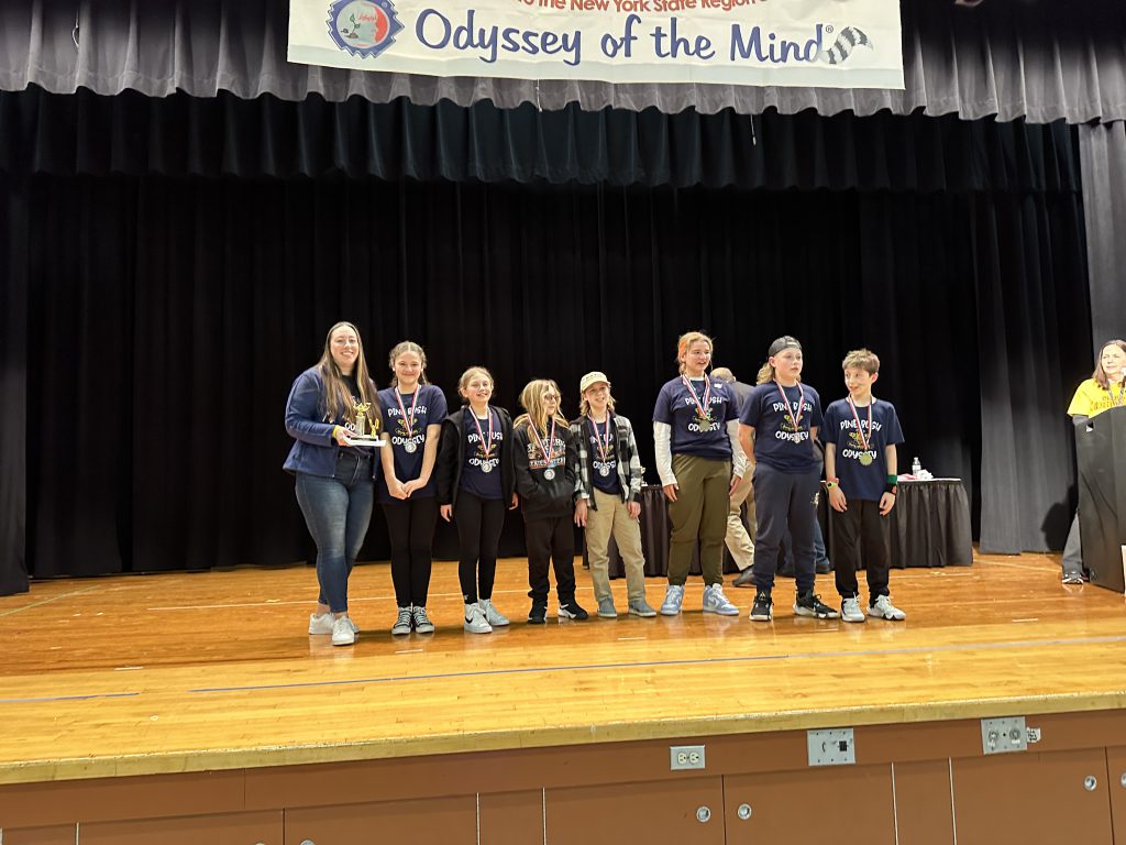 A group of seven middle school age kids on a stage, with a woman on the left. They are all smiling and hae medals around their necks.
