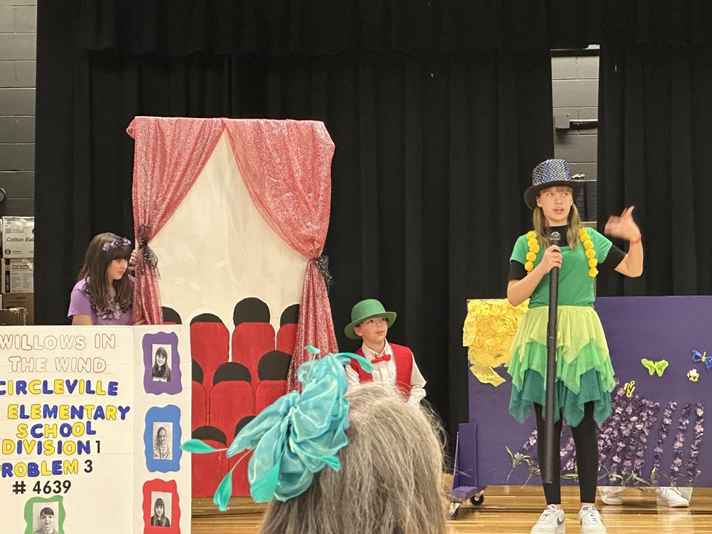 Elementary students in colorful costumes with scenery in the background.