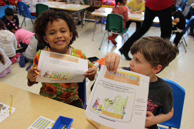 Two kindergarten kids hold up coloring sheets where they drew and colored a garden.
