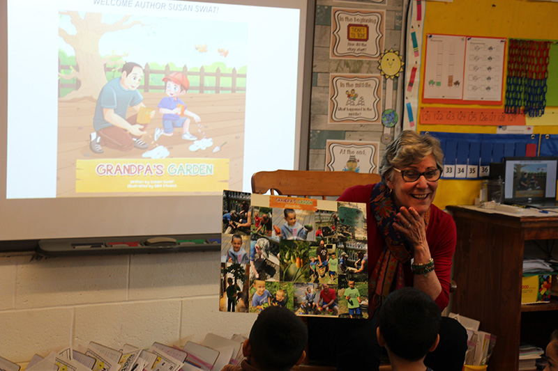 A woman with gray hair holds up a book she is reading to a group of kindergarten kids.