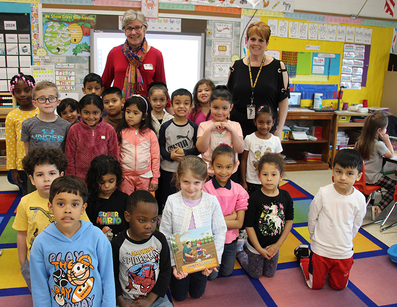 A full class of kindergarten kids sit, and stand with two women. They are smiling.