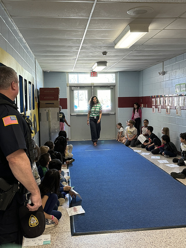 A blue carpet down a hallway with kids sitting on either side of it. A woman stands at the far end of it.