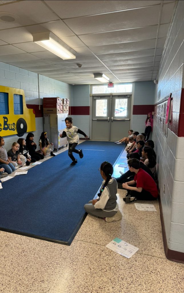 A blue carpet down the center of a hallway with kids sitting on either side of it. An elementary age boy runs down the center of it.