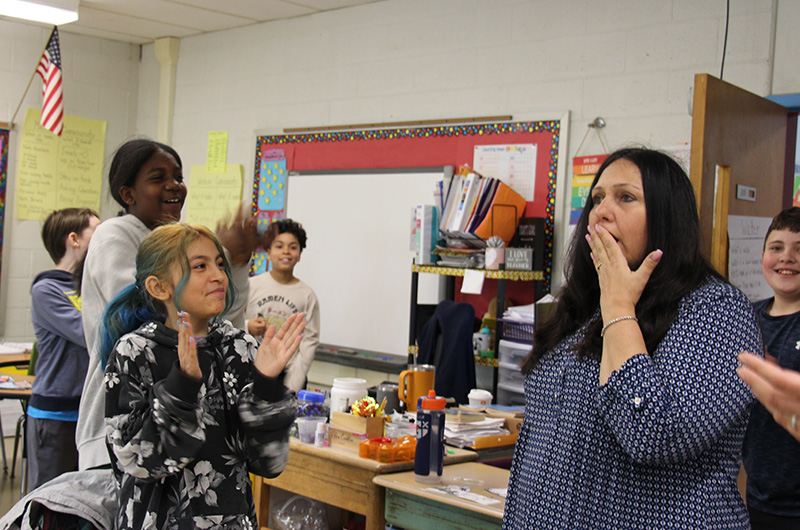 A woman on the right with long dark hair holds her hand up to her mouth in surprise. Fifth-grade students on the left smile and clap.