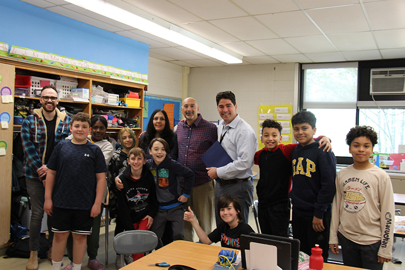 Nine fifth-grade students stand around four adults. All are smiling and have thumbs up.