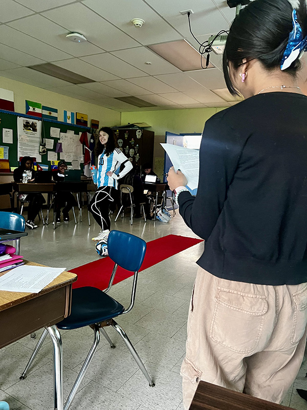 A red carpet in the middle of a classroom with a middle school student walking on it. In the foreground is another student announcing what the other student is wearing.