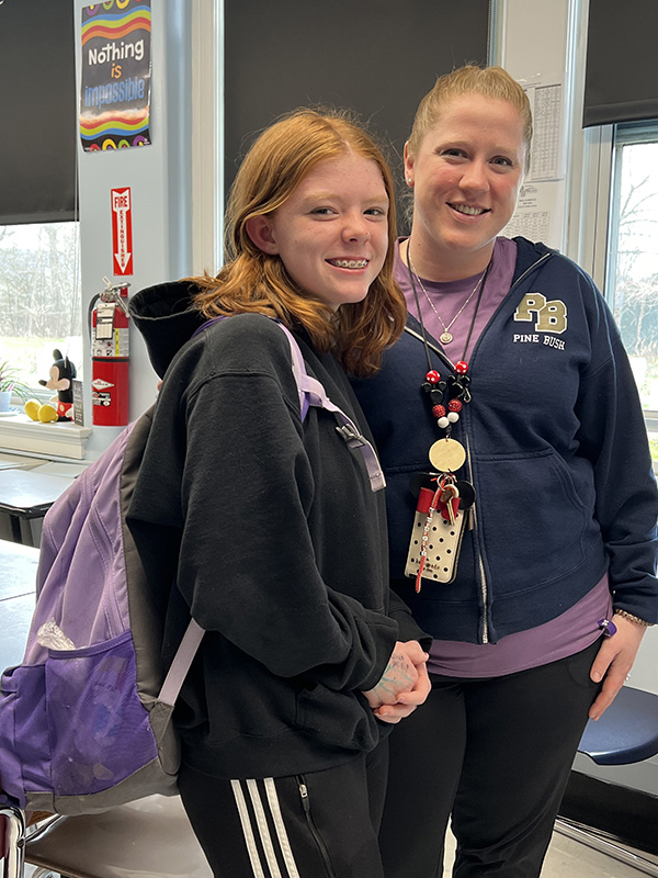 A woman with her hair pulled back on the right, wearing a purple shirt and a middle school girl with red hair, with a purple backpack.