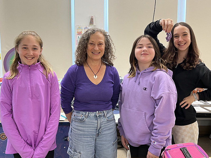 A woman and three middle school girls stand and smile. Three have purple shirts on and one is holding a purple hair tie up.