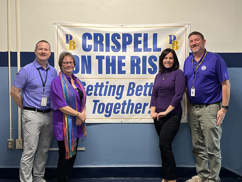 Four adults, two on each side of a sign, stand and smile. All are wearing purple shirts. The sign behind them says Crispell on the Rise Getting Better Together.