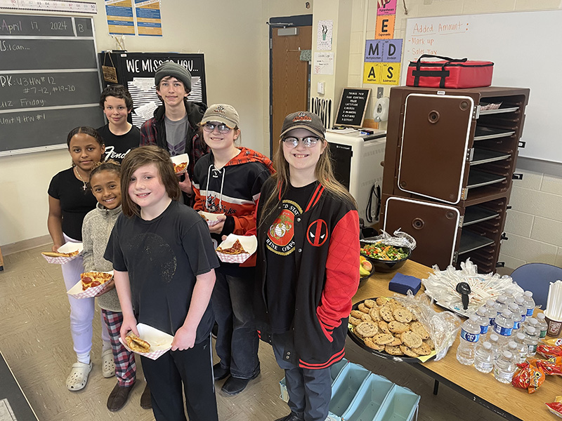 A group of seven middle school kids stand together smiling. They are holding plates with pizza on them and behind them is a table with cookies and salad.