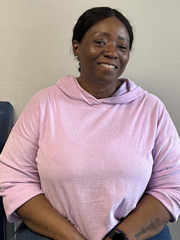 A woman with dark hair pulled back smiles, wearing a light purple shirt.