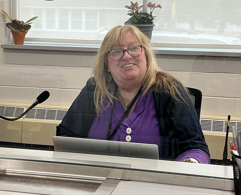 A woman with shoulder-length blonde hair sits at a desk smiling. She is wearing a purple shirt.