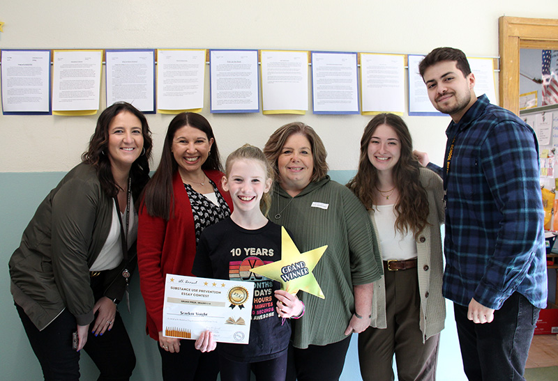 A group of five adults and a young fifth-grade girl who is smiling brightly. She is holding a certificate and a gold star that says Grand Winner.