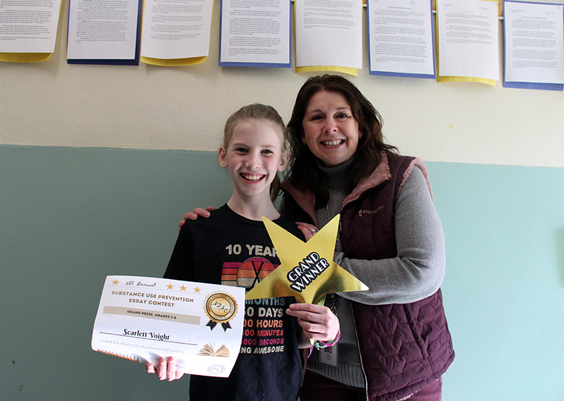 A fifth-grade girl smiles broadly holding a certificate and a large gold star that says Grand Winner. There is a woman standing next to her also smiling.
