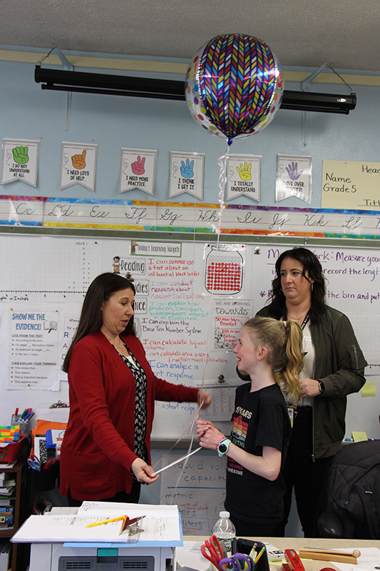 A fifth-grade girl wearing a black shirt and holding a balloon looks at a woman who just gave the balloon to her. There is another woman in the background.