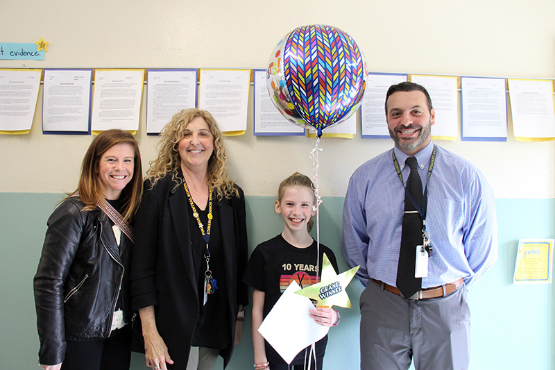 Two women are on the left and a man on the right of a fifth-grade girl holding a large balloon, a certificate and a gold star. They are all smiling.