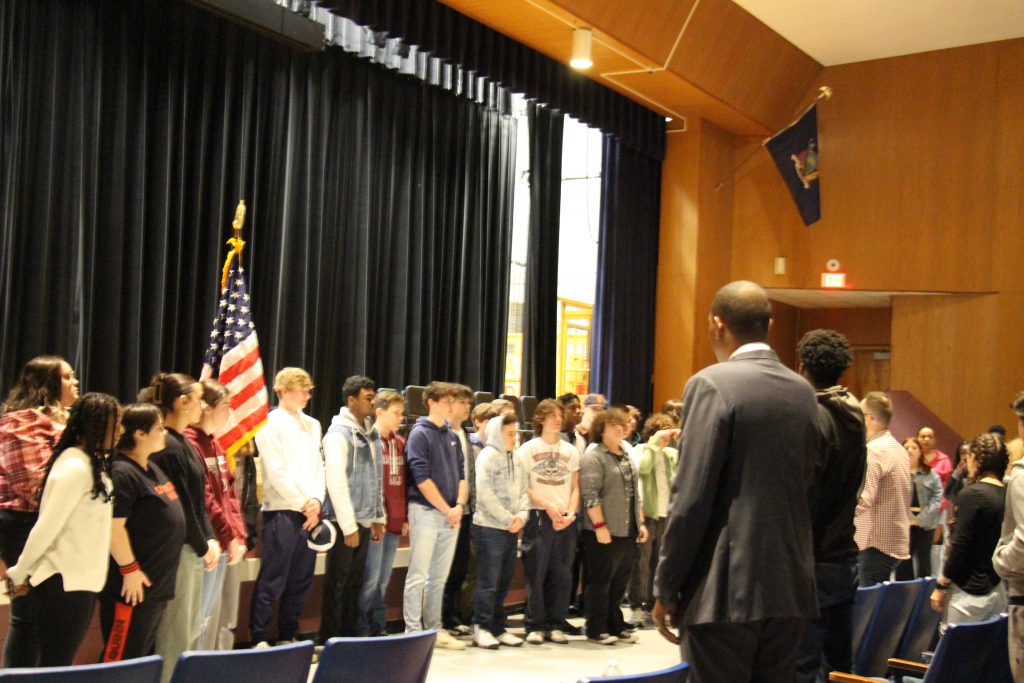 A large numbe rof high school students stand in front of a stage with an American flag behind them. A man stands listening to them sing.