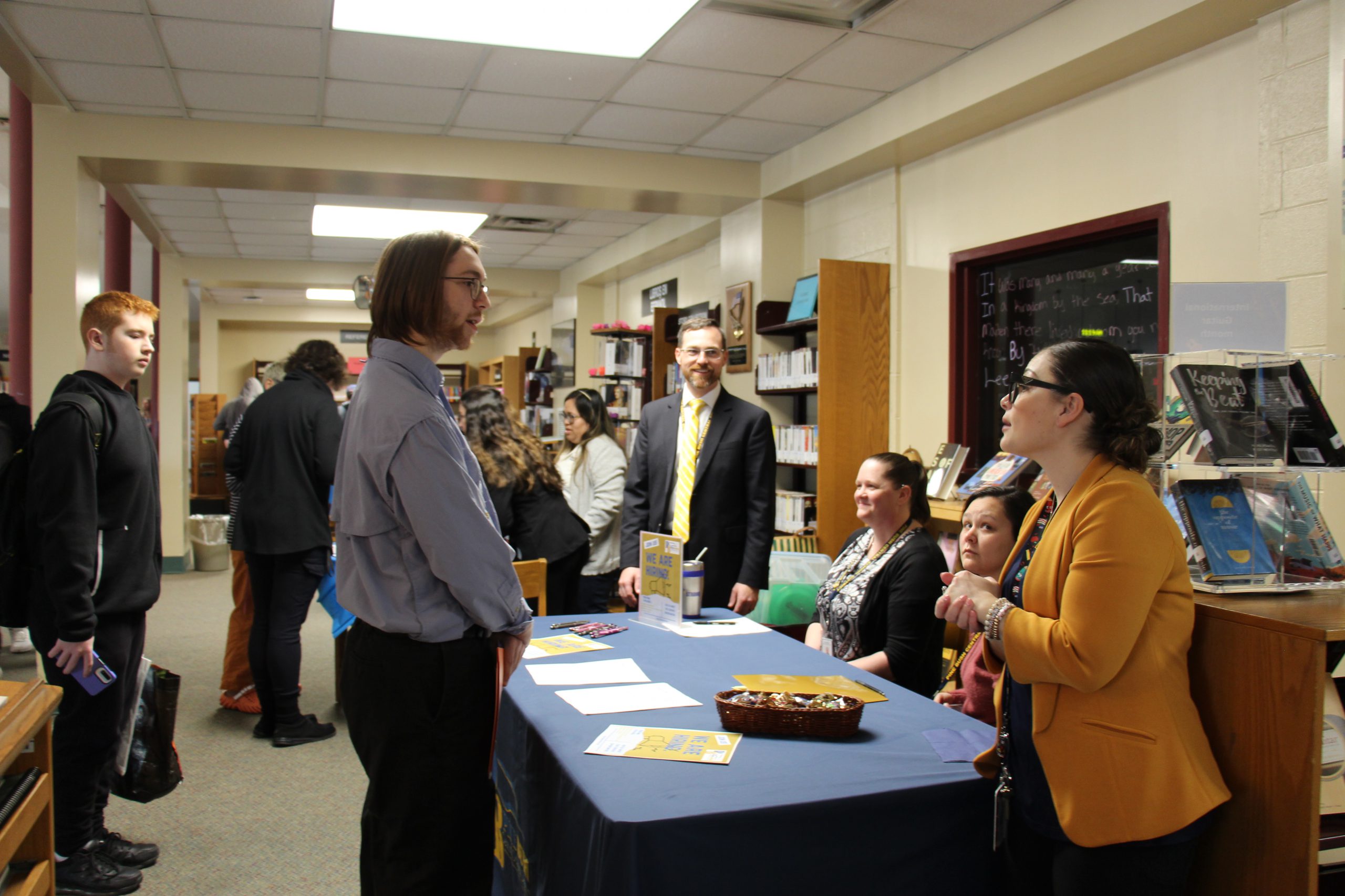 A young man stands at a table covered in a blue tablecloth. A woman in an orange blazer is talking to him and two people are sitting listening. A man is standing and listening.