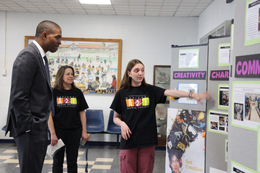 A man in a gray suite listens as a high school student points to artwork on a board and talks about it.