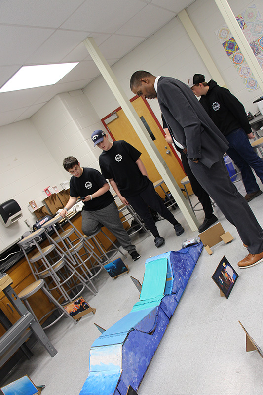 A man in a gray suit looks to the floor where there is a blue roller coaster type track with a car on it. High school students are standing around with remotes.