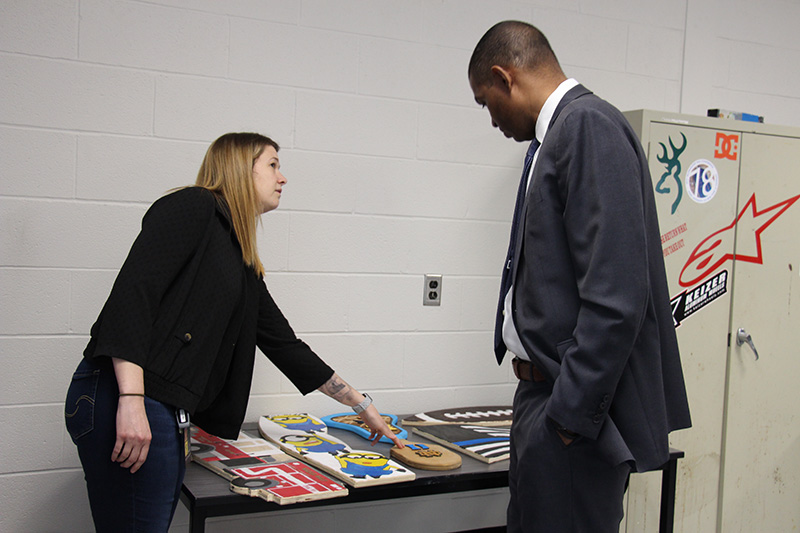 A man in a gray suit looks and listens as a woman with blonde hair points to skateboards her students made.