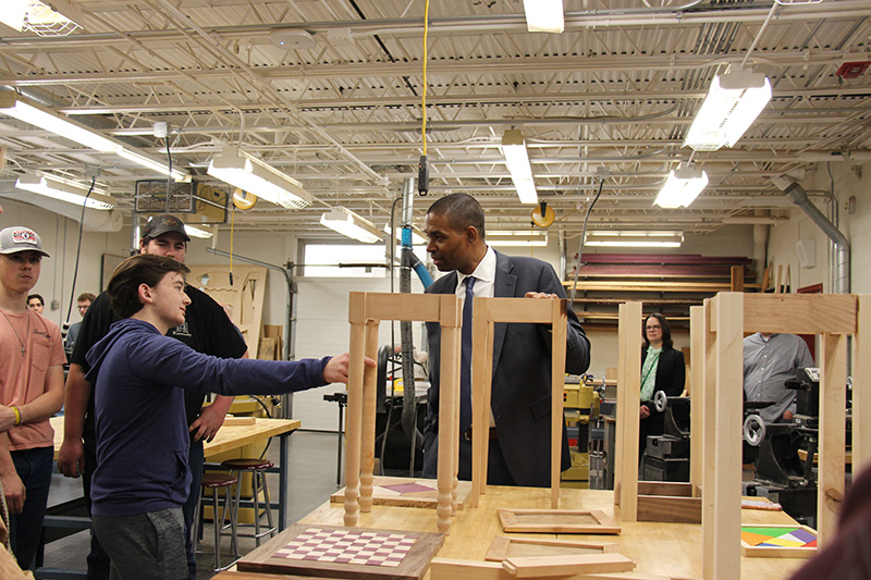A man in a gray suit listens to a high school student describe the table he is making, which is on a bigger table with other projects.