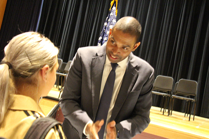 A man in a gray suit talks to a high school student whose hair is pulled back into a pony tail.