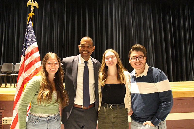 A man in a gray suite stands with three high school students. They are all smiling.