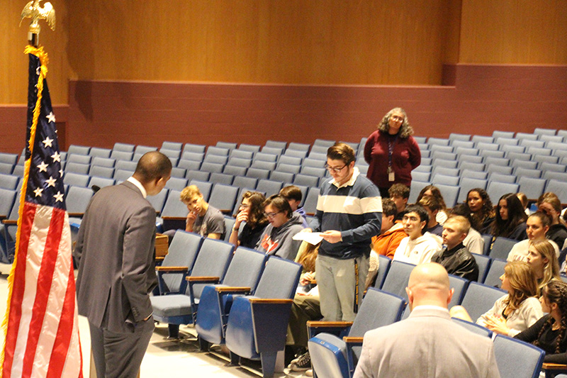 A high school student stands and addresses a man in a gray suit. They are in an auditorium with many other people.