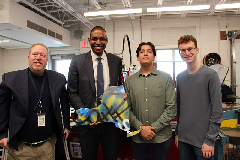 Two men on the left stand with two high school boys and are holding a metal replica of the state of New York.