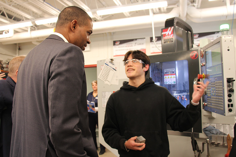A high school student in a black sweatshirt points to a large machine and explains to a man in a gray suit what is happening.