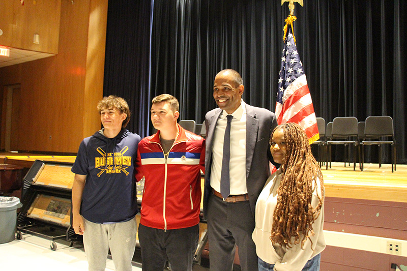 A man and three high school students stand together in front of an American flag smiling.