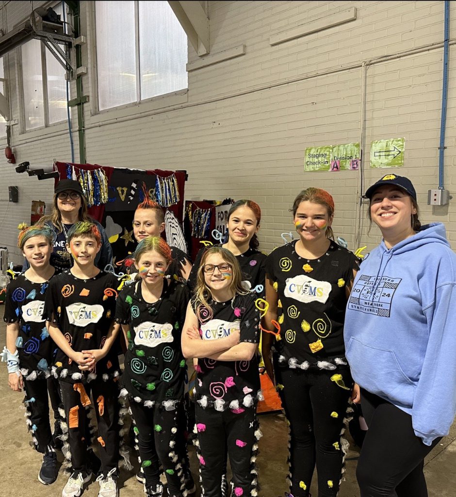 Two adults in light blue sweatshirts stand at either end of a group of seven middle school students dressed in black outfits, each with a different color cutouts on the outfit. They are all smiling.