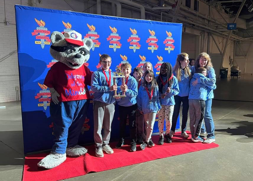 A person in a large racoon costume wearing a shirt that says Odyssey of the Mind, stands with a group of seven elementary students and two women. Two of the students are holding a large trophy. All are smiling.