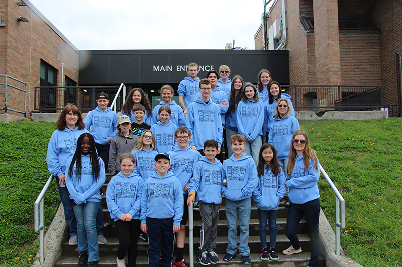 A group of 25 people - five adults and 20 elementary and secondary students - stand on the steps to a building. They are all wearing light blue  hoodies that say Pine Bush Odyssey 2024 New York State Finals