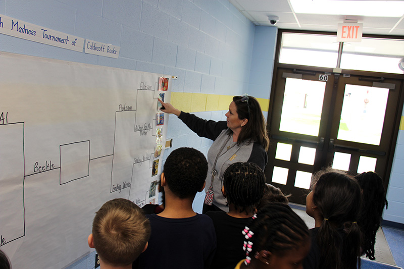 A woman with shoulder length dark hair points to a bracket written on poster paper hanging on the wall as several young elementary students watch.