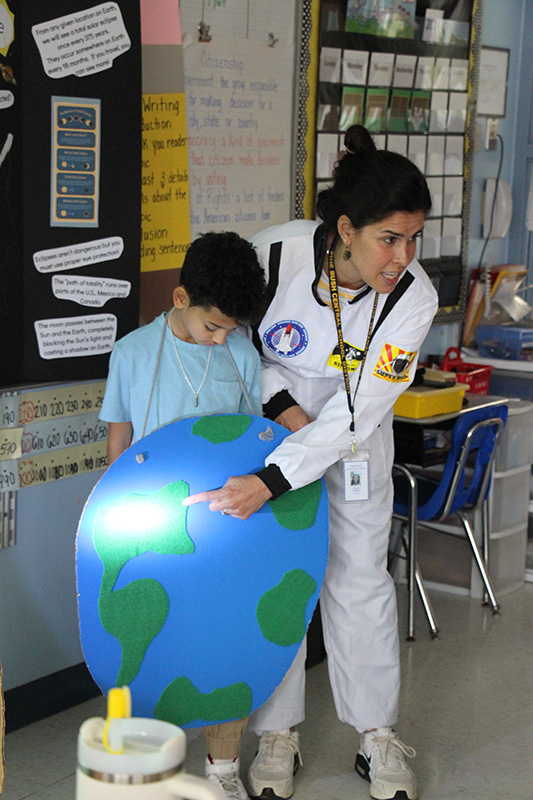 A woman in a white space suit points to a light reflected on a big cutout of the earth that is hanging on a second grade student.