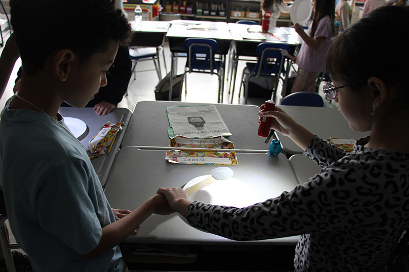 A darkened classroom. Two elementary students use a flashlight and a small cut out moon to show a reflection on a desk.