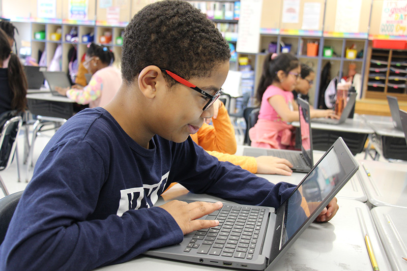 An elementary age boy with glasses and short dark hair, wearing a blue long-sleeve shirt works on his Chromebook.