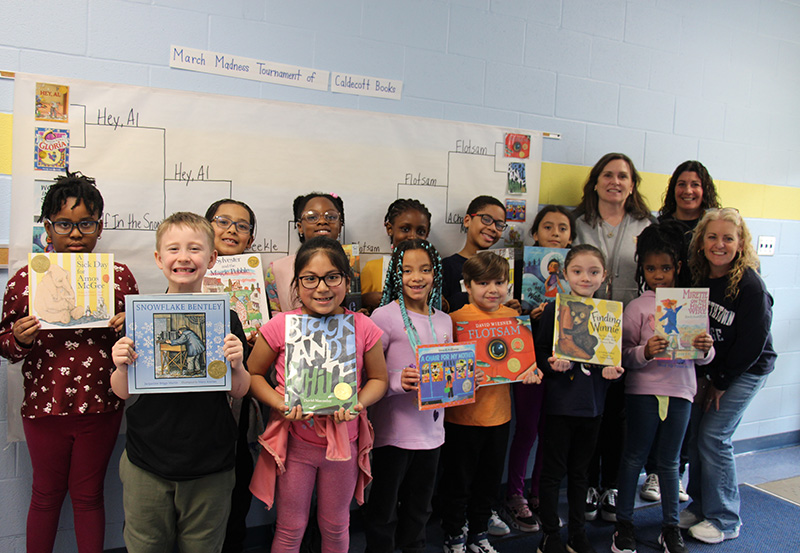 An entire second-grade class holds up books with three of their teachers.