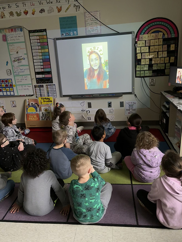 A group of young elementary students sitting on the floor listening to a woman in a video.