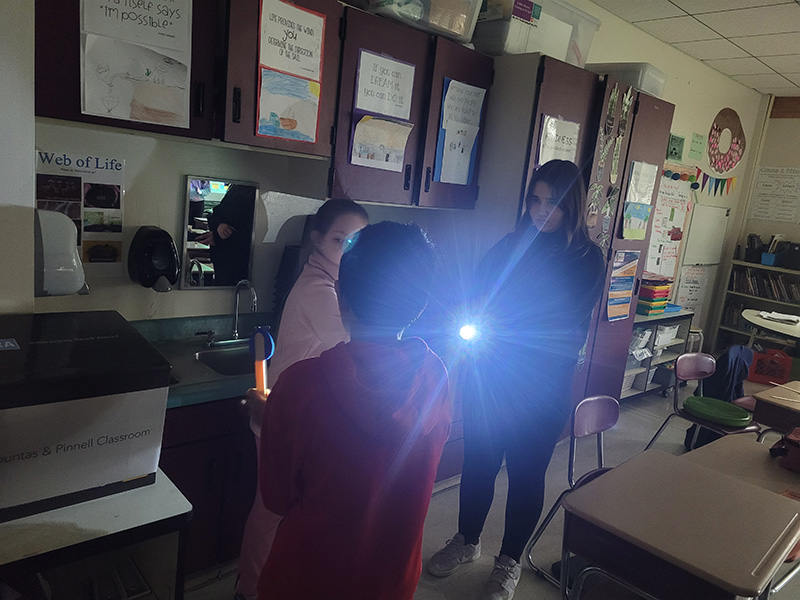 Students in a darkened room use a flashlight and cardboard to show how a shadow is cast like will be done during the eclipse.