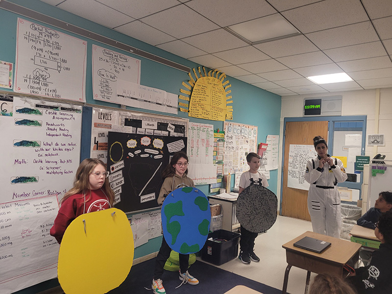 Three students stand in front of a classroom. One has a larger yellow sun, another has an earth, another has a moon. A woman in a white astronaut suit stands to the side.