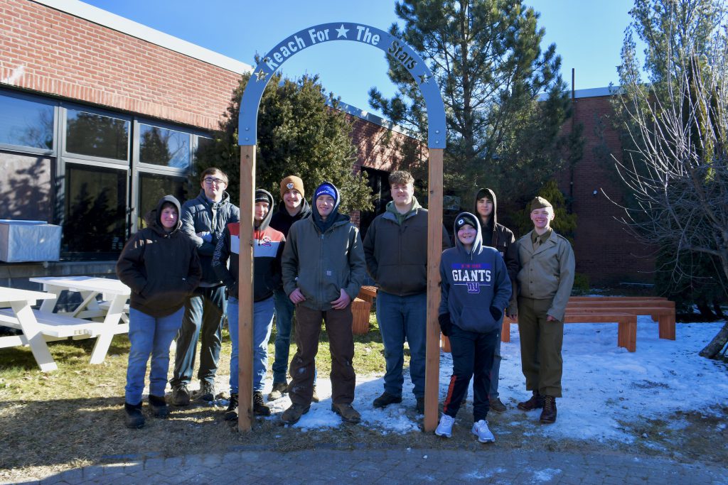 A group of high school kids stand under an archway build from wood and metal. It says Reach for the Stars.